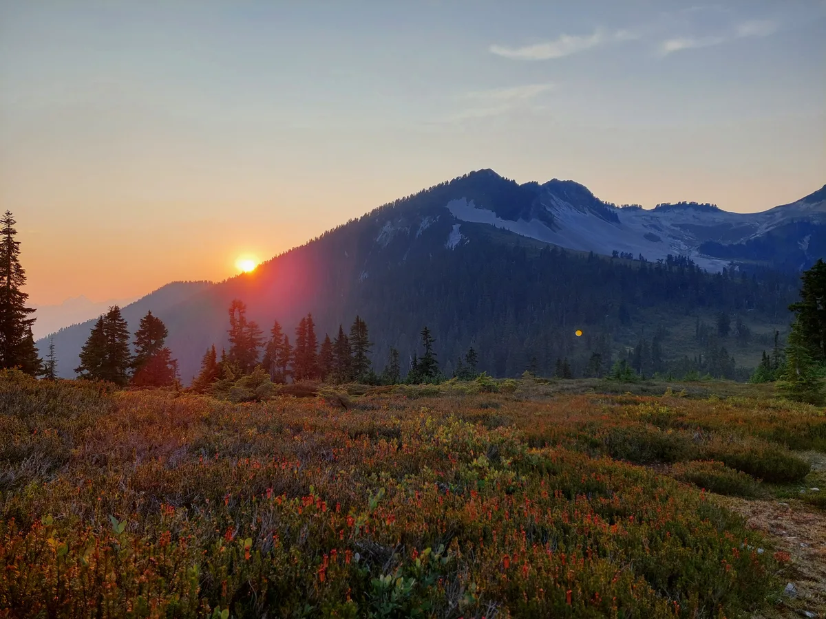 The setting sun peeking past a mountain ridge above an alpine meadow