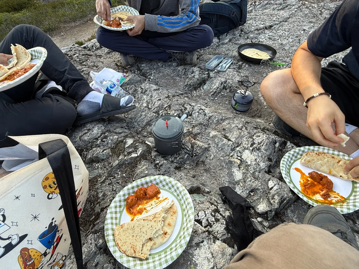 A group sharing a meal on rocks beside a camping stove