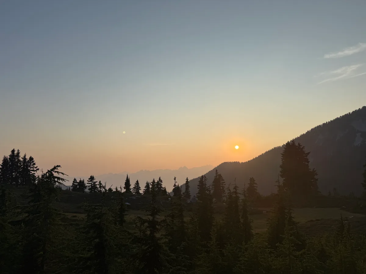 An orange sun above silhouetted mountains and evergreen trees