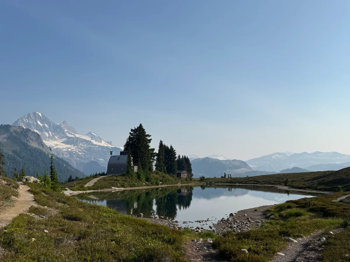 A cabin and evergreens reflected in a lake beneath snowy mountains
