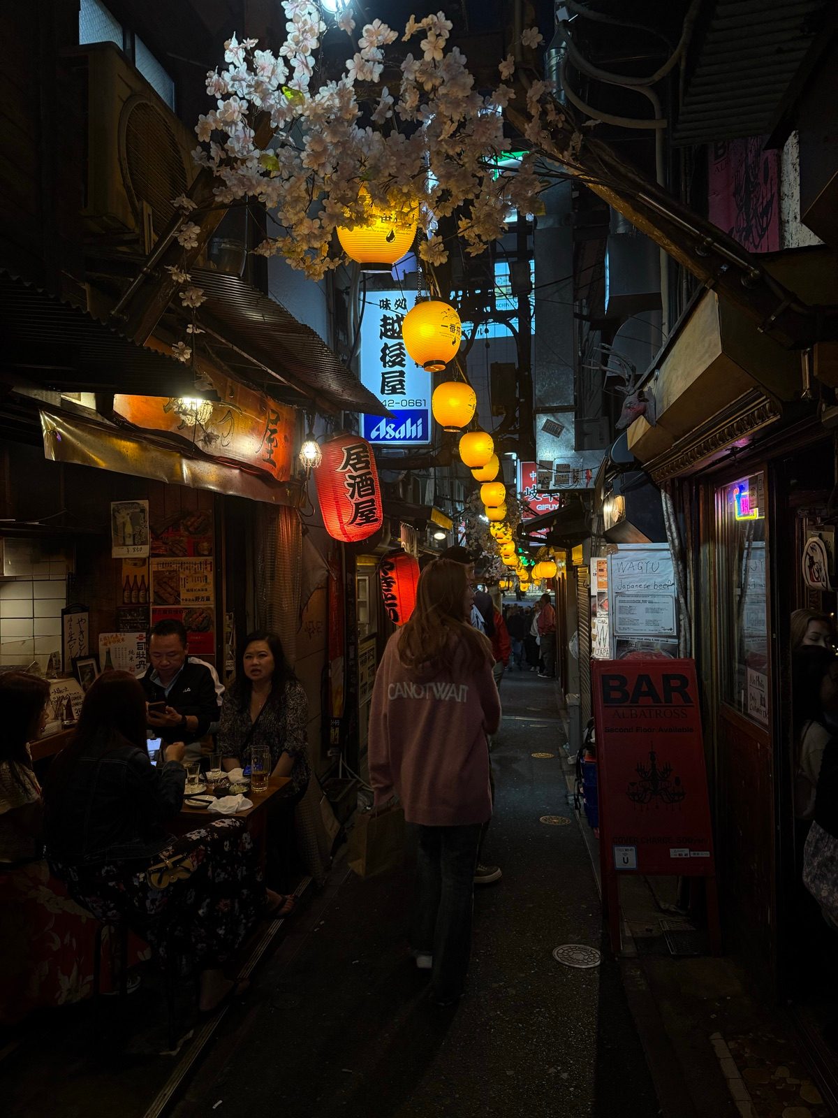 A narrow restaurant alley with glowing lanterns and overhead blossoms