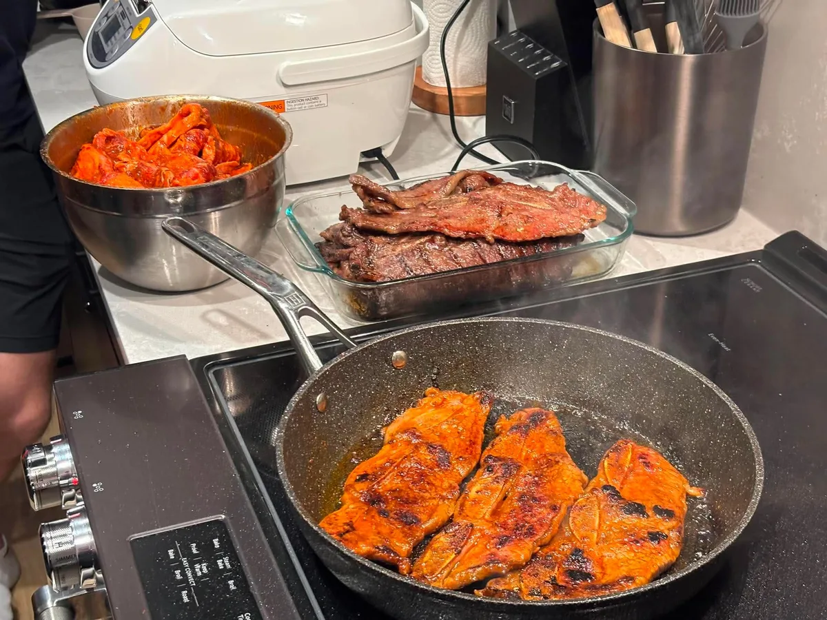 Food cooking in a skillet beside trays and pots on a kitchen counter